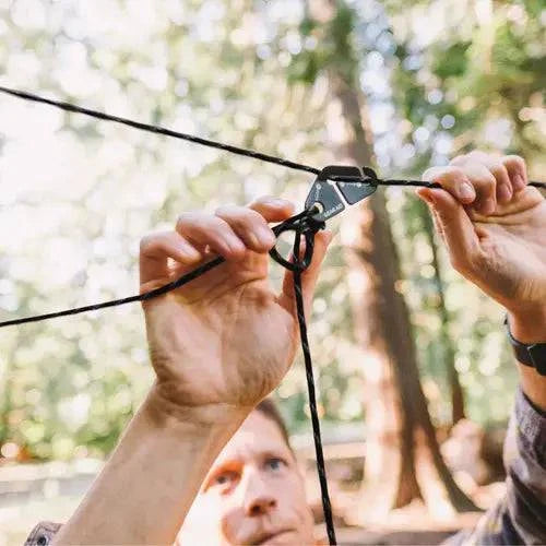 A person’s hands use the Gear Aid Taut Line Kit to secure 550 paracord between trees outdoors, with a blurred forest background. The image highlights the hands and the Gear Aid device.