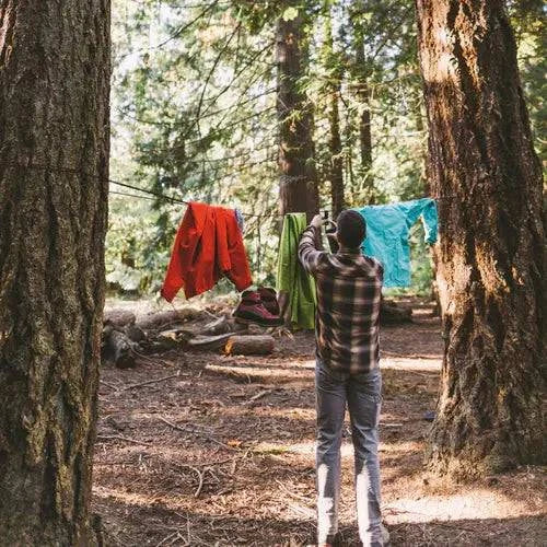A person stands in a sunlit forest, photographing colorful clothes hung on a line between two large trees using the Gear Aid Taut Line Kit with 550 paracord and line tensioners.