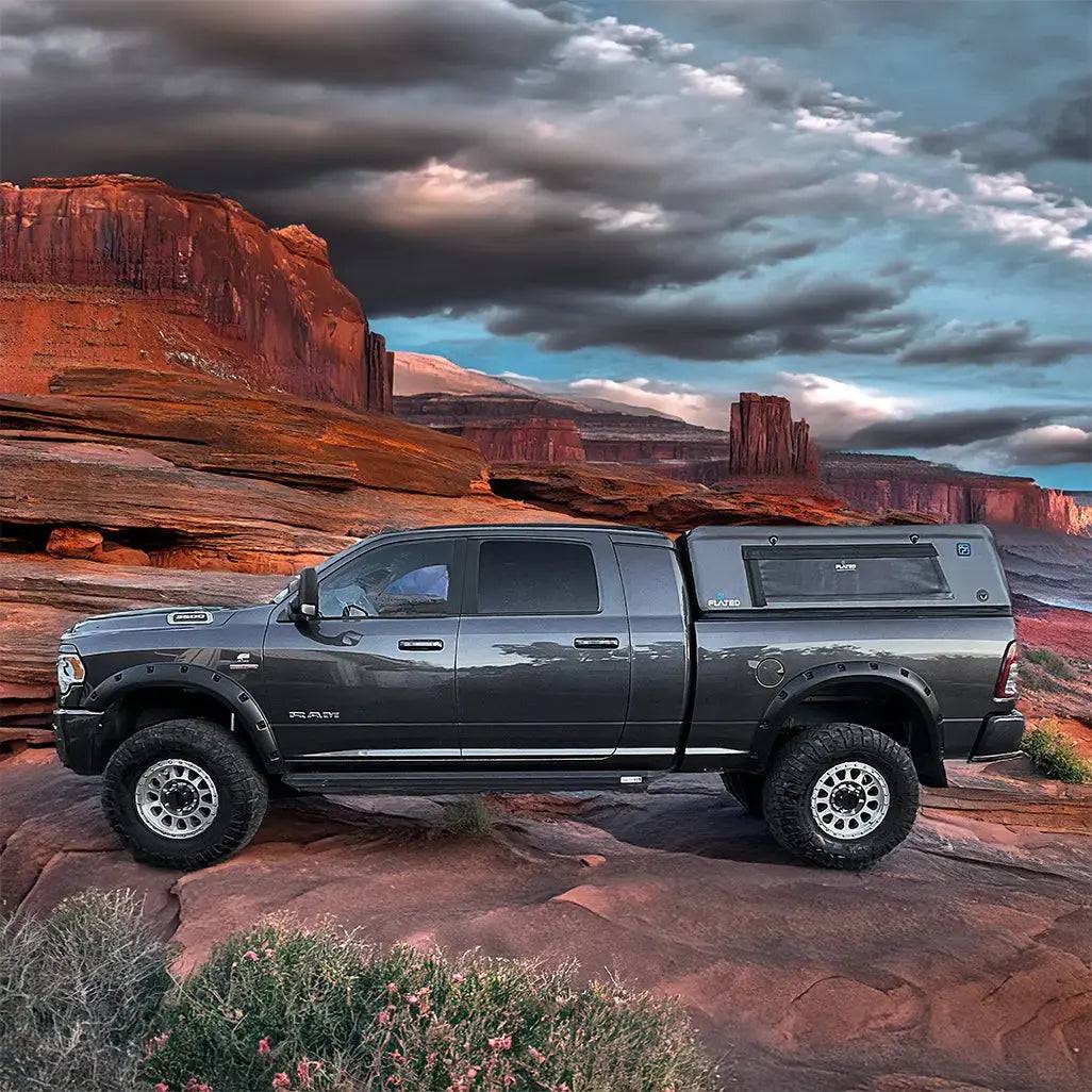 A FLATED FULL-STANDARD DT CAP64— a lightweight truck shell— sits on a black pickup with a full-size bed, parked amid rocky terrain beneath dramatic sunset clouds and surrounded by striking red rock cliffs.