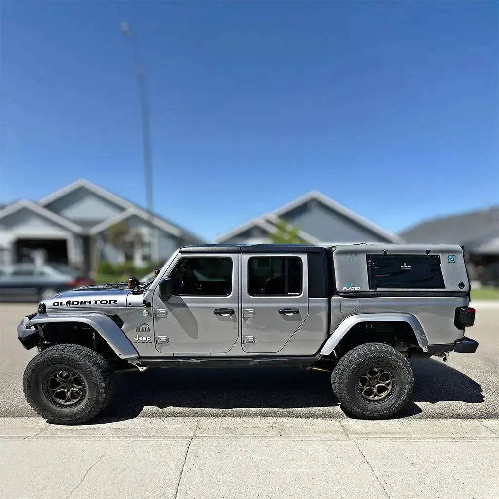 A silver Jeep Gladiator with large off-road tires and a FLATED MID-SHORT CAP51 inflatable truck shell is parked on a suburban street in front of modern houses under a clear blue sky.