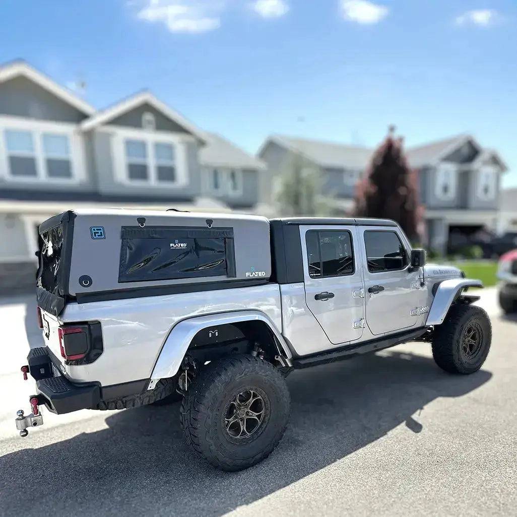 A silver Jeep Gladiator with large off-road tires and a black FLATED MID-SHORT CAP51 topper is parked on a suburban street in front of modern two-story houses on a sunny day.