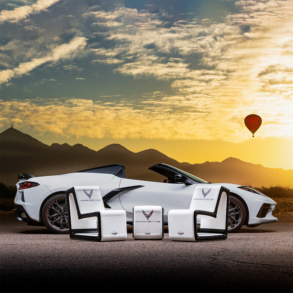 At sunset with mountains behind, a white convertible sports car is parked. In front, FLATED's COMPACT-SIZE CORVETTE SET offers four portable inflatable seats for frunk-gating, while a hot air balloon floats above.