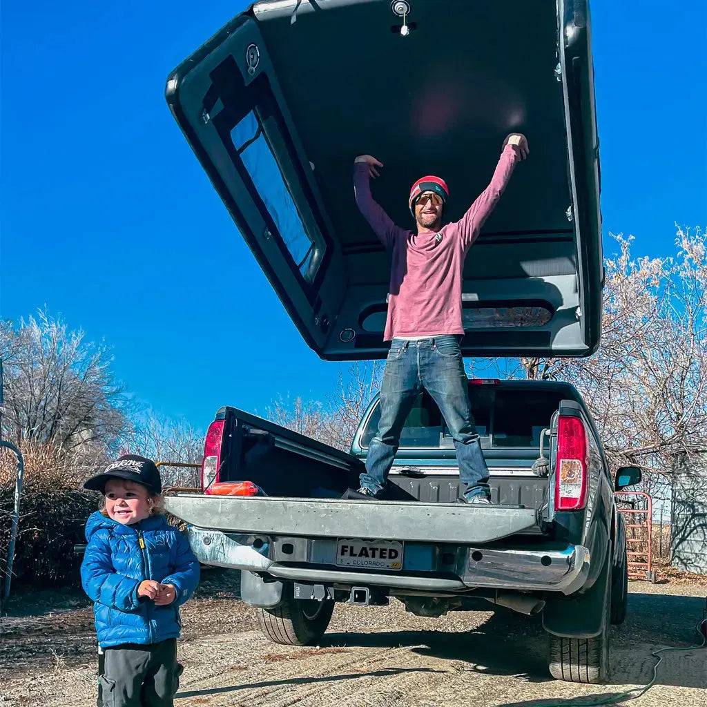 A man stands in the bed of a FLATED MID-LONG CAP60 truck with the topper open, arms raised. A smiling child in a blue jacket stands nearby under a clear blue sky, with trees in the background.