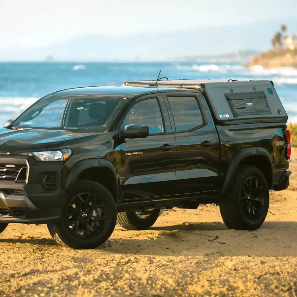 A black Toyota Tacoma fitted with a FLATED MID-SHORT CAP50 truck cap is parked on sandy ground by the ocean, with waves and a distant coastline in the background.