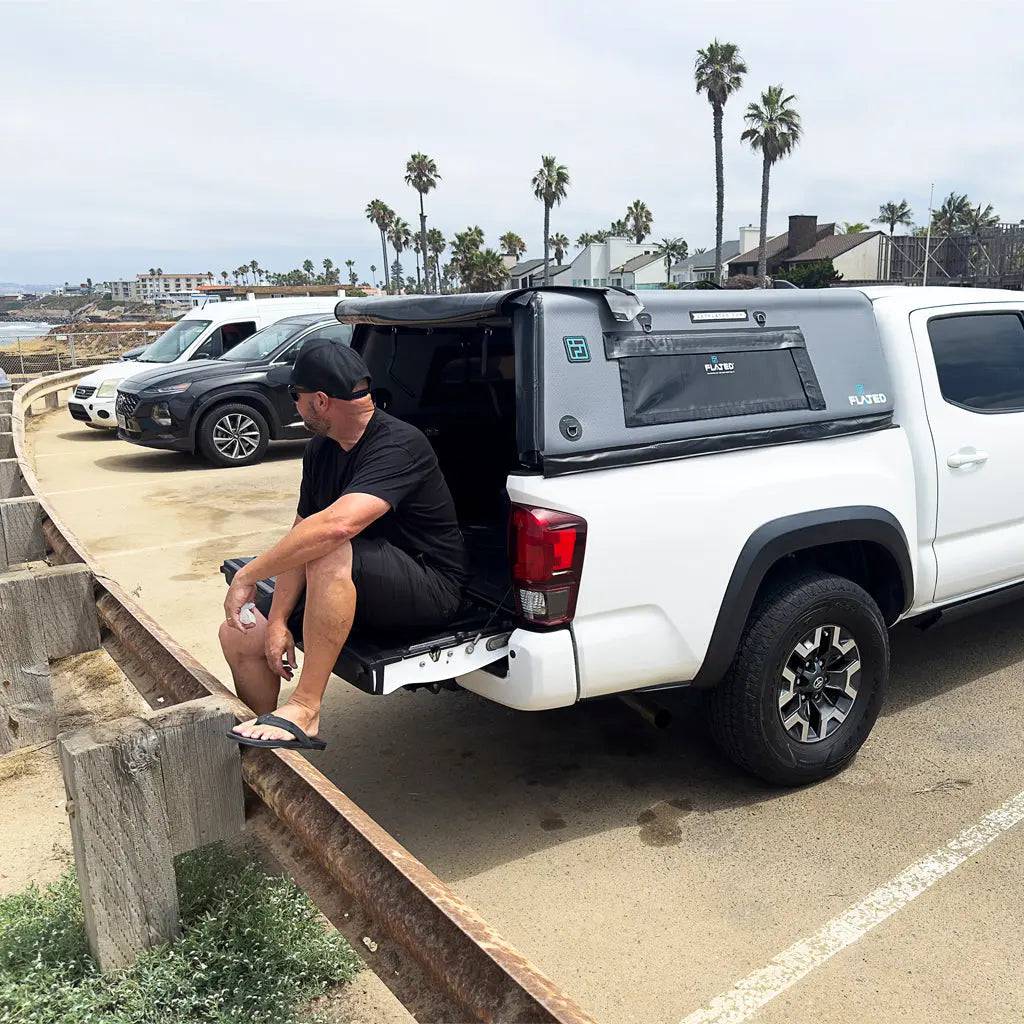A man in black clothes and flip-flops sits on the tailgate of a white Toyota Tacoma equipped with the FLATED MID-SHORT CAP50, parked by the beach with palm trees, cars, and buildings under a cloudy sky in the background.