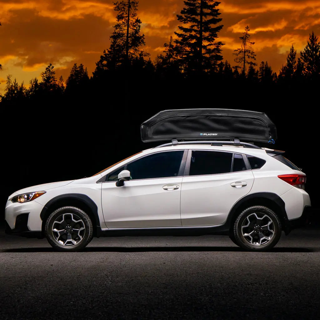A white SUV with a FLATED SPORT CARRIER15 (OPEN BOX) rooftop cargo carrier is parked on a road, silhouetted against a dramatic orange sunset and tall pine trees—an ideal setup for travel adventures.