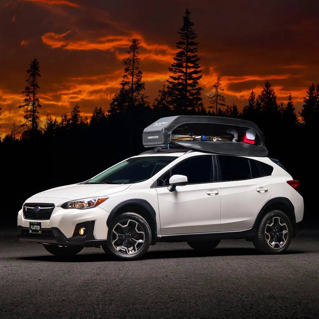 A white SUV with a FLATED SPORT CARRIER15 (OPEN BOX) rooftop cargo box is parked on a road, outlined by evergreen trees and an orange sunset—ideal gear storage for all your travel adventures.
