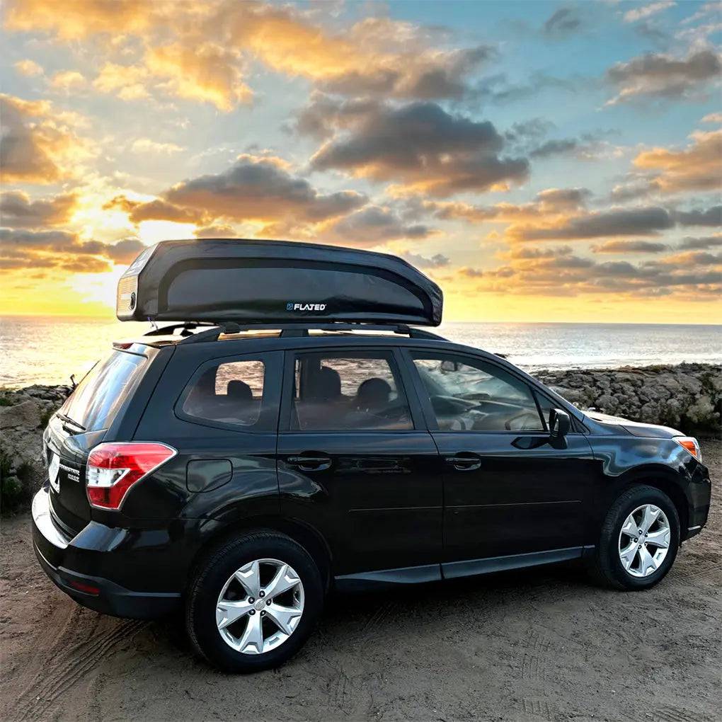 A black SUV with a FLATED SPORT CARRIER15 (OPEN BOX) rooftop cargo carrier is parked by a rocky shoreline at sunset, offering ample gear storage for travel adventures with the ocean and vibrant clouds in the background.