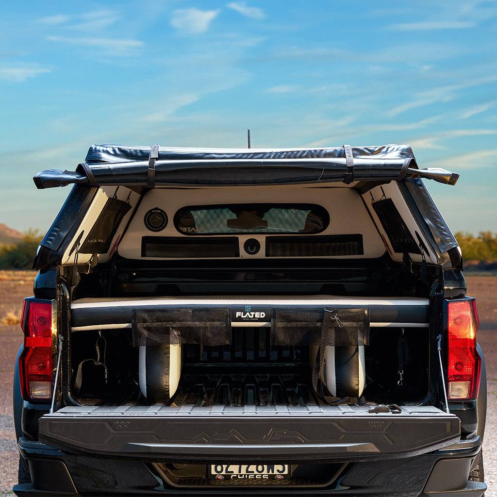 A black GM pickup with an open tailgate displays surfboards and gear on the FLATED FULL-STANDARD DECK66 SILVERADO air mattress, parked outdoors under a clear blue sky.