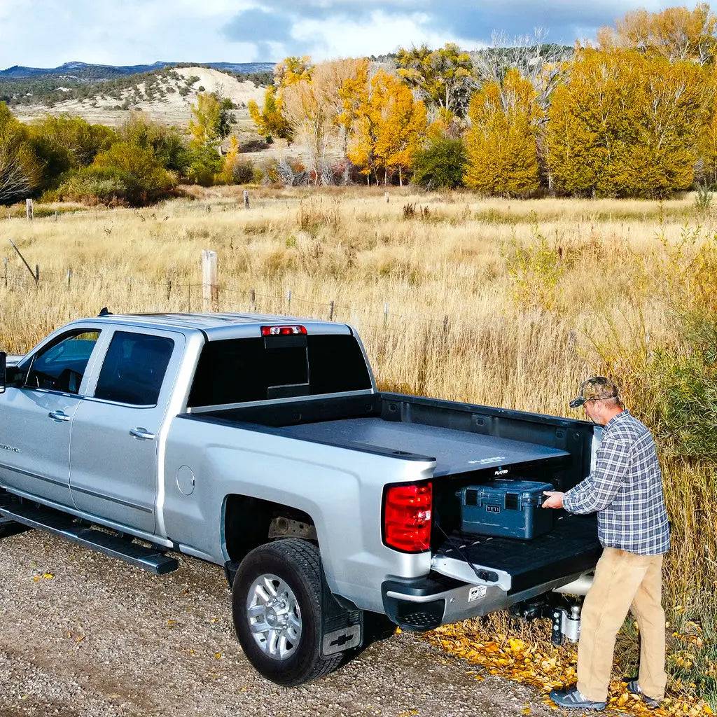 A man loads a toolbox into the bed of his silver pickup, equipped with the FLATED MID-SHORT DECK50 (OPEN BOX), parked on a gravel road with autumn hills and trees in the background.
