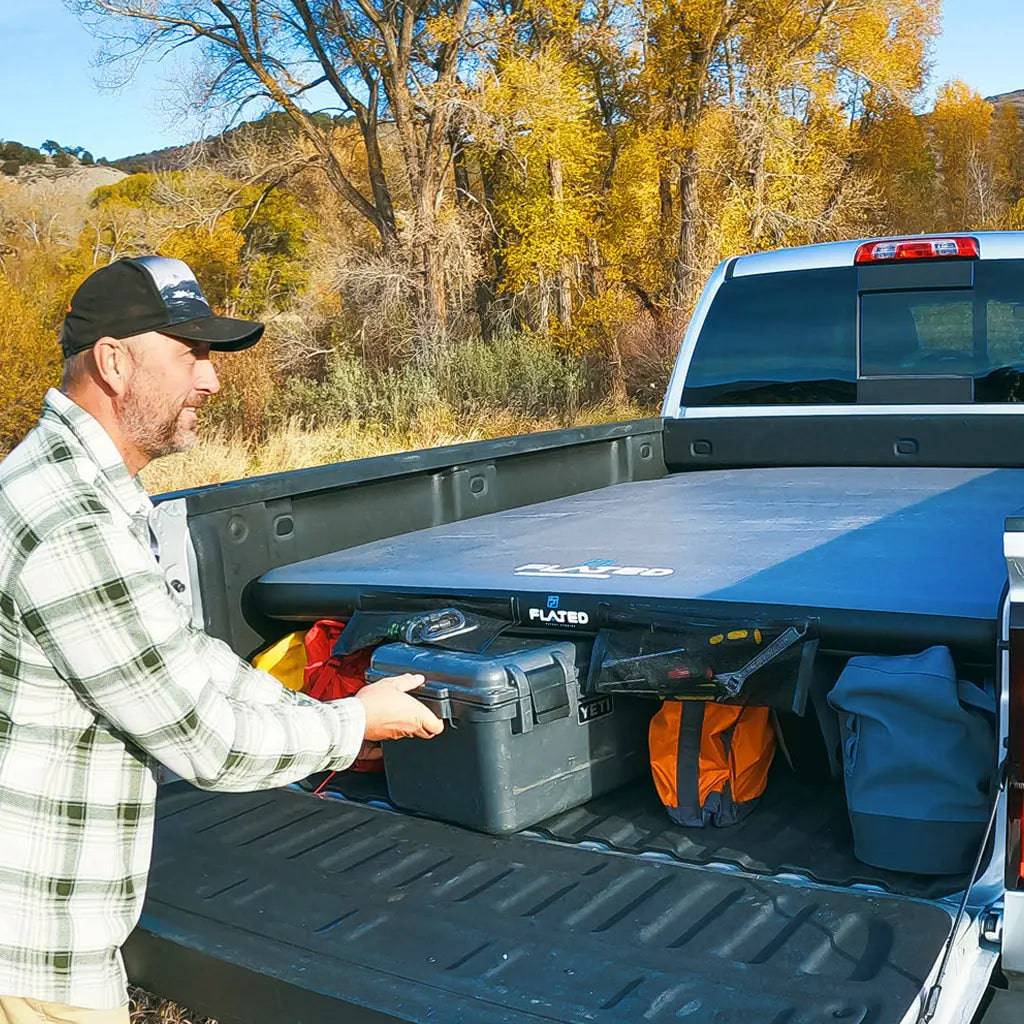 A man in a plaid shirt and cap stands by his adventure truck, its open bed packed with gear and bags, featuring a FLATED FULL-SHORT DECK56 inflatable truck bed platform, all set against autumn trees and a clear sky.
