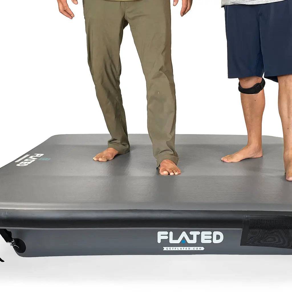 Two people stand barefoot on a large, dark gray FLATED FULL-SHORT DECK56 inflatable truck bed platform. One wears khaki pants, the other navy shorts and a knee brace. The white background highlights this versatile adventure rig.