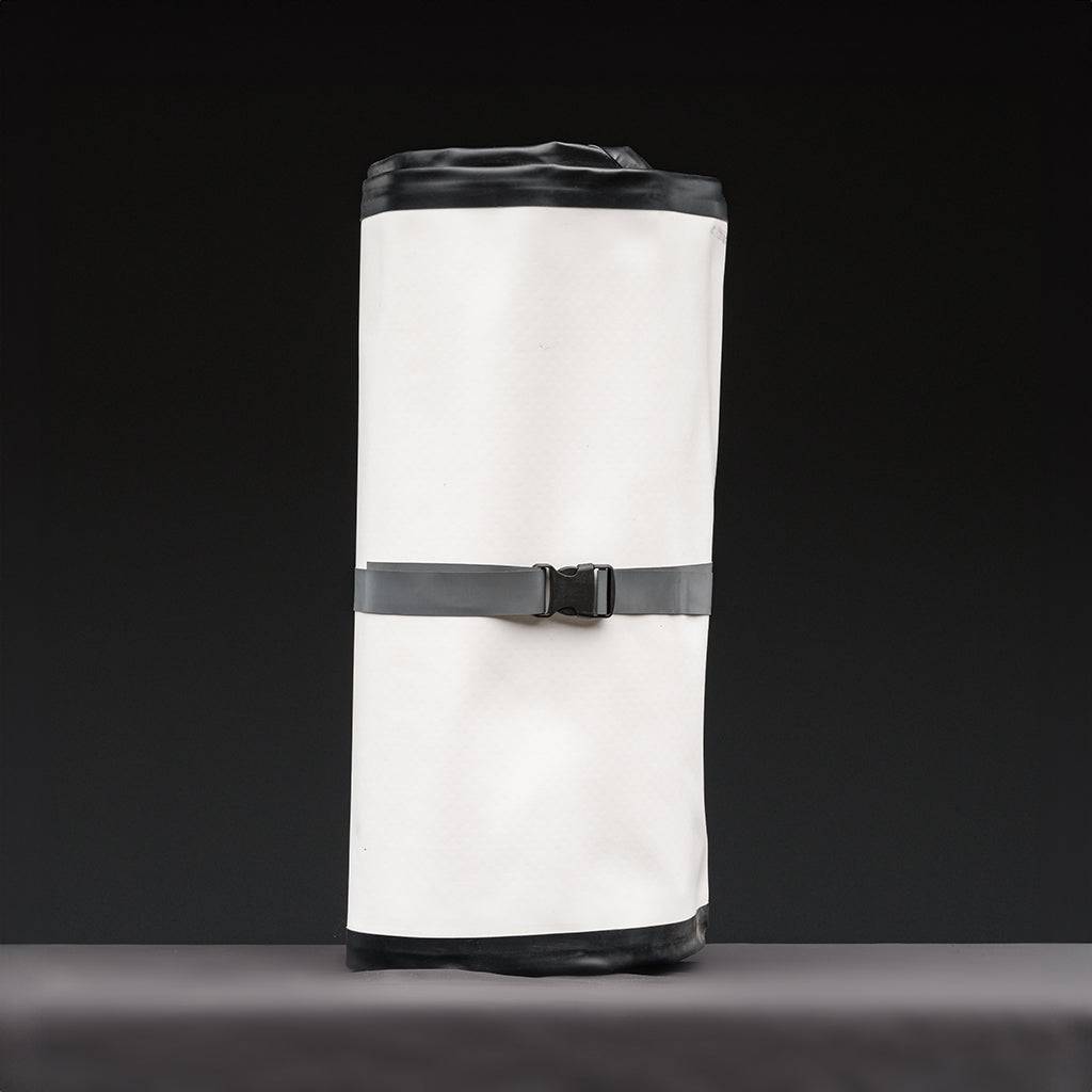 A FLATED MID-SIZE TABLE60 nylon camping table bag, white and black, stands upright on a gray surface against a dark background, secured at the middle with a black strap and buckle.