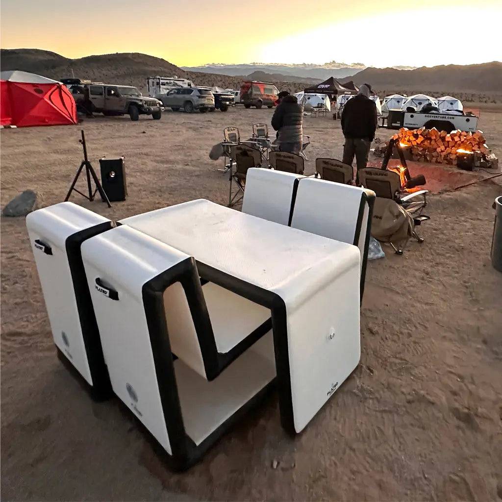 The FLATED MID-SIZE TABLE60, a modern minimalist white camping table with black edges, is set up on sandy ground at a campsite where people gather around a campfire with tents and vehicles at sunset.