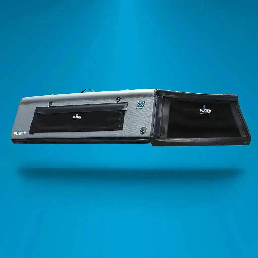 A large, rectangular black FLATED FULL-SHORT CAP54 (OPEN BOX) rooftop cargo carrier with a windowed side pocket is shown against a solid blue background, featuring the FLATED brand name on the main section and pocket.