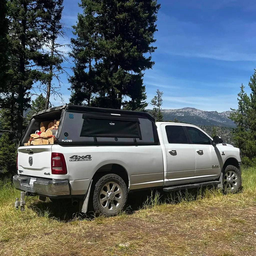 A white pickup truck equipped with the FLATED FULL-LONG MR80 truck topper is parked on grass near tall pines, its full-size bed loaded with firewood, with mountains in the background beneath a blue sky.