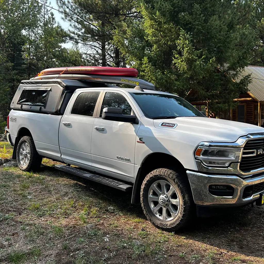 A white RAM pickup with a FLATED FULL-LONG MR80 topper is parked on grass by trees, paddleboards and gear secured on its roof. A cabin stands in the pine trees in the background.