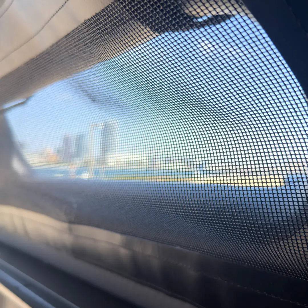 View through a mesh screen shows blurred city buildings and blue sky, with a window frame and a FLATED FULL-STANDARD MR66 inflatable truck shell for a full-size truck bed in the foreground.