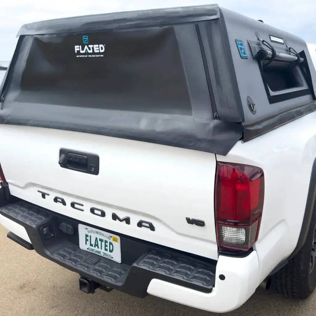A white Toyota Tacoma mid-size long bed truck features a black FLATED MID-LONG CAP60 inflatable topper covering the bed. The license plate reads “FLATED.” The truck is parked outdoors on a paved surface.
