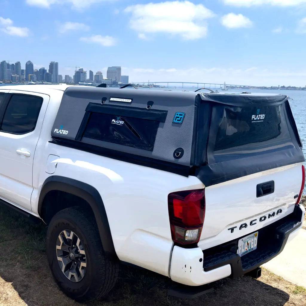 A white Toyota Tacoma with a FLATED MID-SHORT CAP50 (black) is parked on grass by water, with a city skyline and bridge in the background beneath a partly cloudy sky.