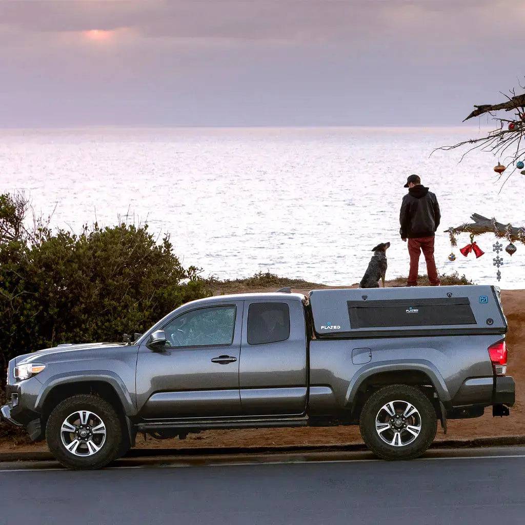 A person and a dog stand on a cliff at sunset near a gray FLATED MID-LONG CAP60 truck, with a small decorated tree to the right.