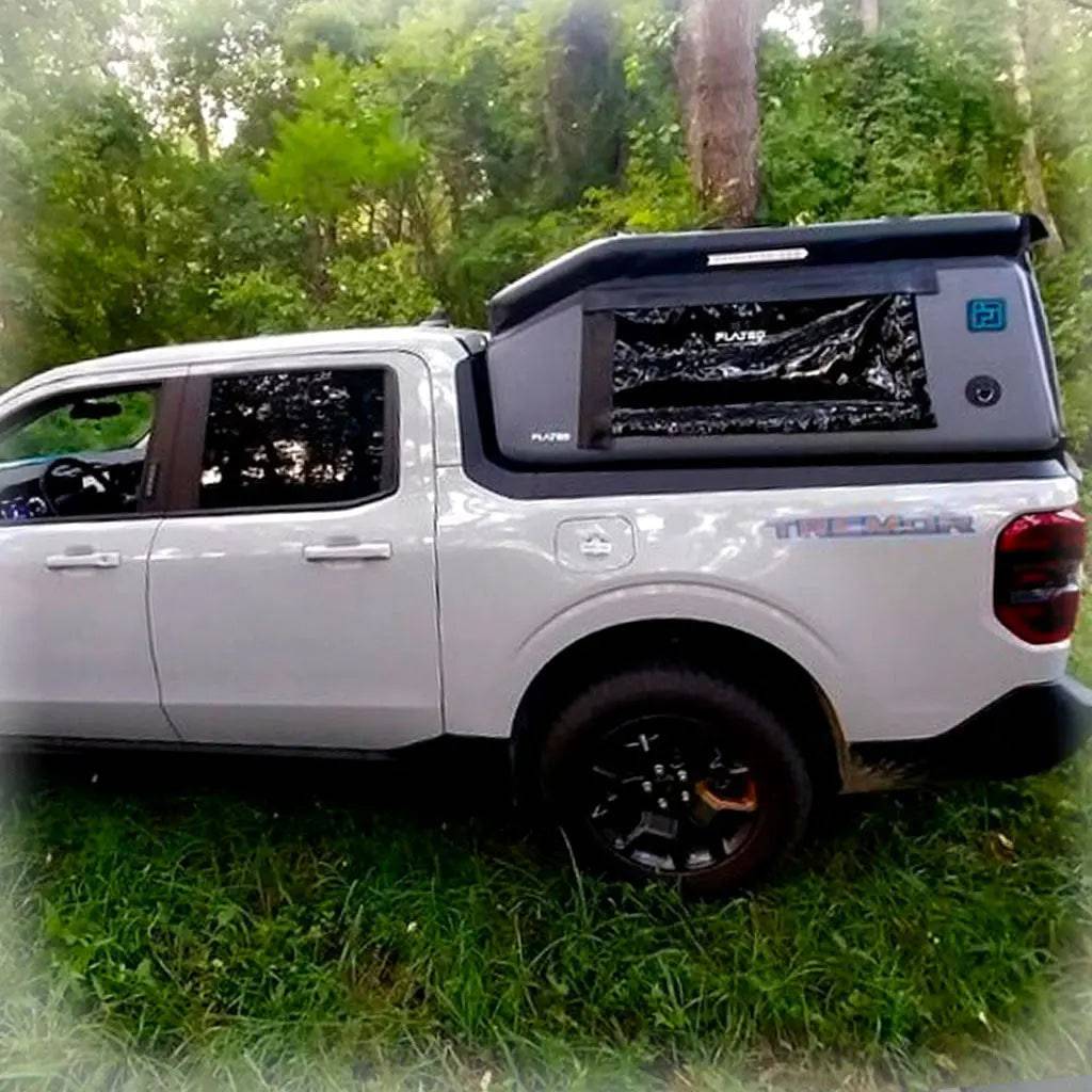 A FLATED COMPACT-MAVERICK MR46 black truck topper is installed on a white pickup parked on grass in a wooded area. The topper's roof is popped up, with trees and green foliage visible in the background.
