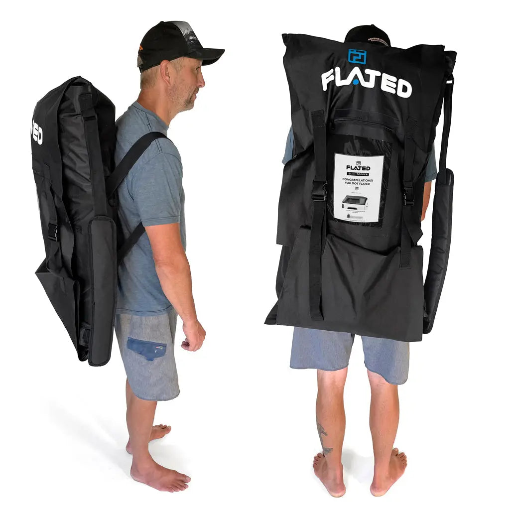 A barefoot man in a cap, t-shirt, and shorts carries a large FLATED MID-SHORT DECK50 (OPEN BOX) bag—taller than his torso—on his back. The black bag features shoulder straps for easy carrying of your inflatable truck bed platform.