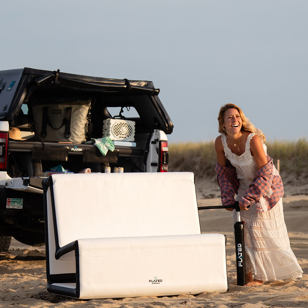 A woman in a white dress and plaid shirt smiles as she inflates a FLATED MID-SIZE LOVESEAT (OPEN BOX) by a parked vehicle on a sandy beach, with the setting sun lighting up the grass-covered dunes behind her.