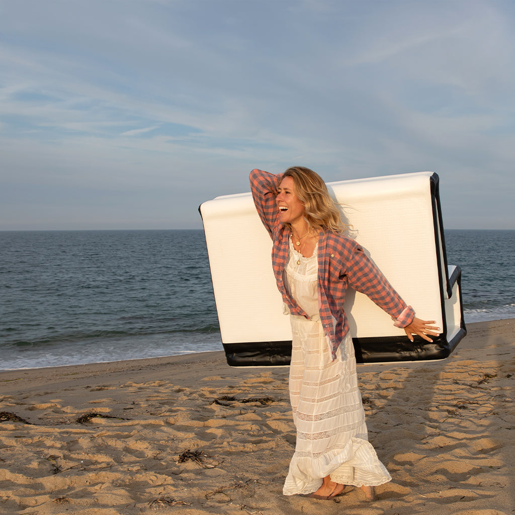 A woman wearing a white dress and plaid shirt smiles while carrying the FLATED MID-SIZE LOVESEAT (OPEN BOX) on her back, walking barefoot along a sandy beach by the ocean.