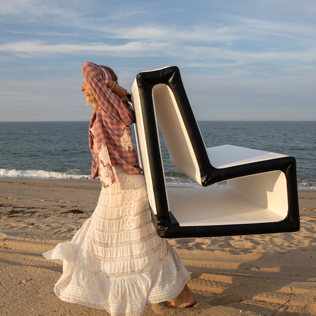 A woman in a plaid shirt and long white skirt walks barefoot on the beach, carrying a FLATED MID-SIZE LOVESEAT (OPEN BOX) on her back, with the ocean and sky in the background.