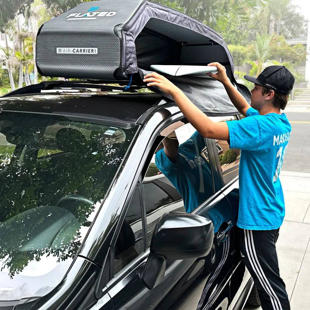 Wearing a blue shirt and cap, a person installs the FLATED SPORT CARRIER15 (OPEN BOX) atop a black car on a suburban street, adding extra gear storage for travel adventures amid houses and trees.