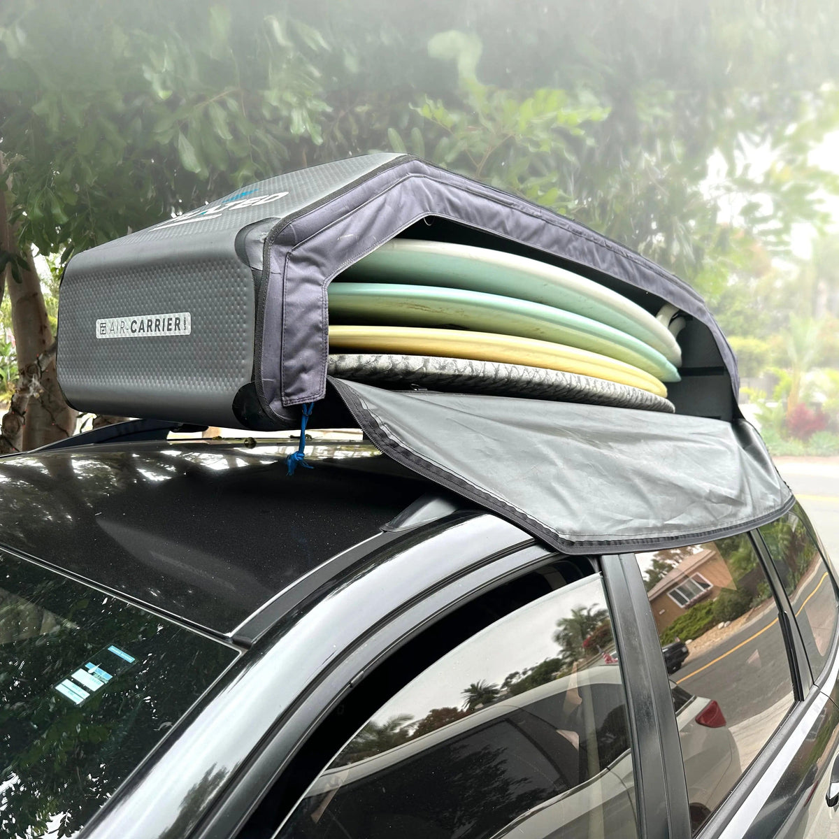 A black car with an open FLATED SPORT CARRIER15 (OPEN BOX) rooftop cargo carrier holds stacked surfboards, making gear storage easy. Trees and a residential street appear in the background, setting the scene for travel adventures.