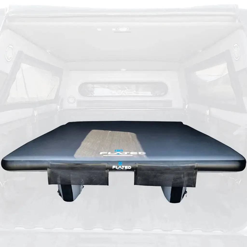 The FLATED MID-SHORT DECK50 (OPEN BOX), a black surfboard with two fins, wood-textured detail, and PLATZED logo, rests in an inflatable truck bed shown with a translucent effect—perfect for mobile adventure rigs.