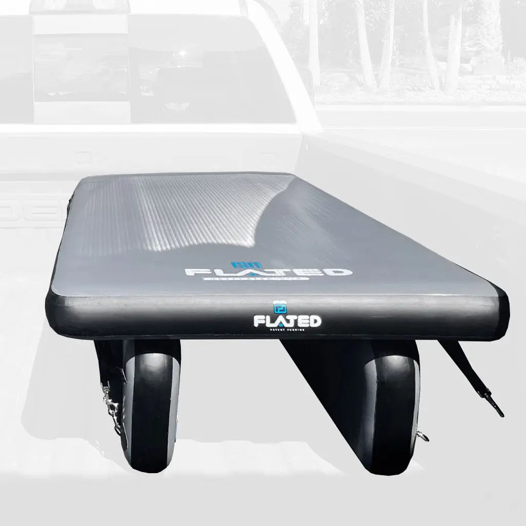 A black FLATED COT DECK60 inflatable sleeping platform with pontoons rests in a white pickup truck bed, set against a backdrop of the adventure rig and trees, showcasing this innovative Air-Deck Cot.