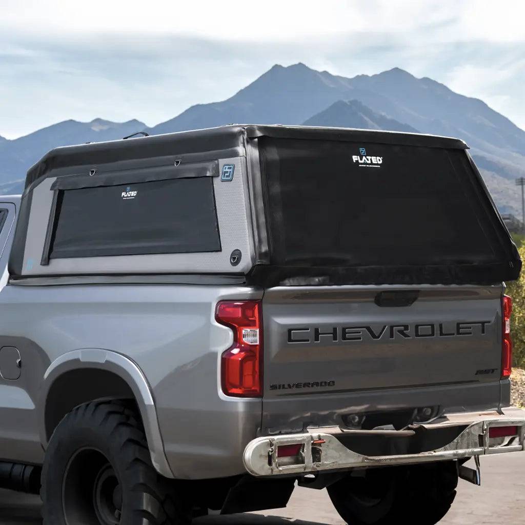 A silver Chevrolet Silverado with a black FLATED FULL-STANDARD MR66 inflatable shell covers its truck bed, parked outdoors with mountains in the background beneath a partly cloudy sky.