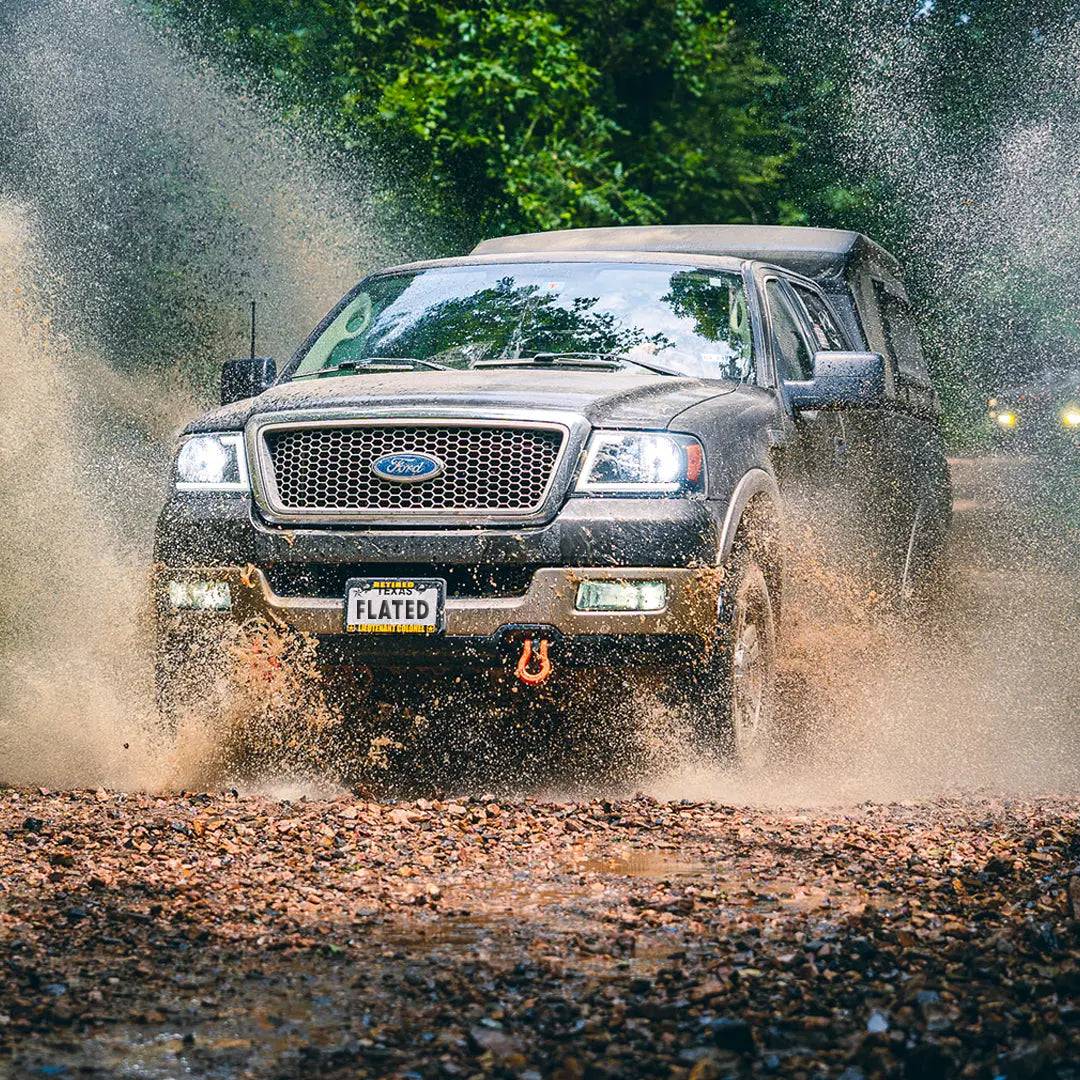 A black Ford SUV with a FLATED FULL-LONG MR80 inflatable truck shell and FLATED license plate splashes through a muddy puddle on a rocky dirt road, water flying, framed by green trees in the background.