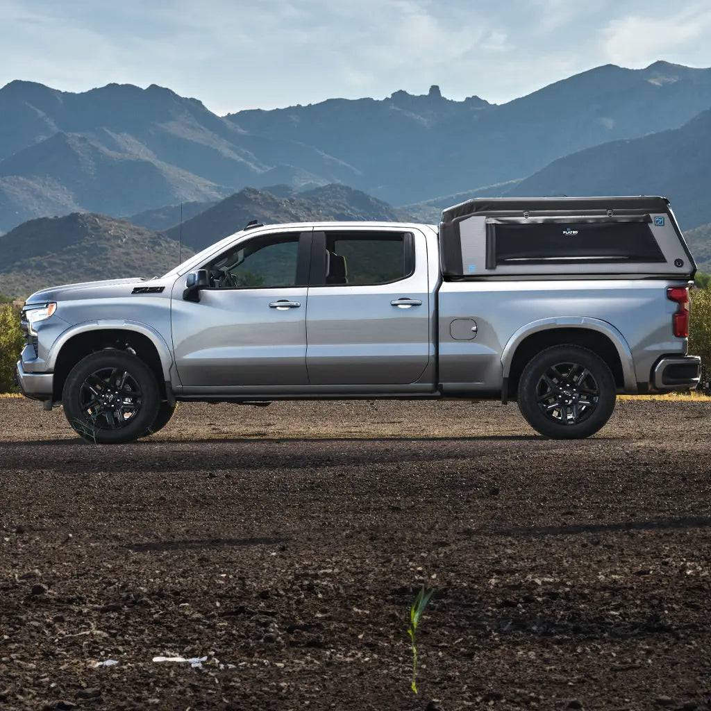A FLATED FULL-STANDARD MR66 truck topper covers the silver pickup's full-size bed as it sits on a gravel road, with mountains and a partly cloudy sky in the background.