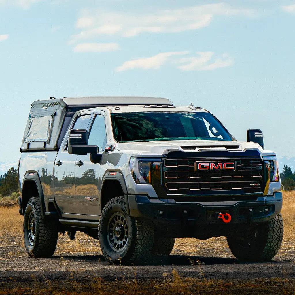 A silver GMC pickup fitted with off-road tires and a FLATED FULL-STANDARD MR66 truck topper is parked on dry grassland under a partly cloudy sky, with the front grille showcasing the red GMC logo.