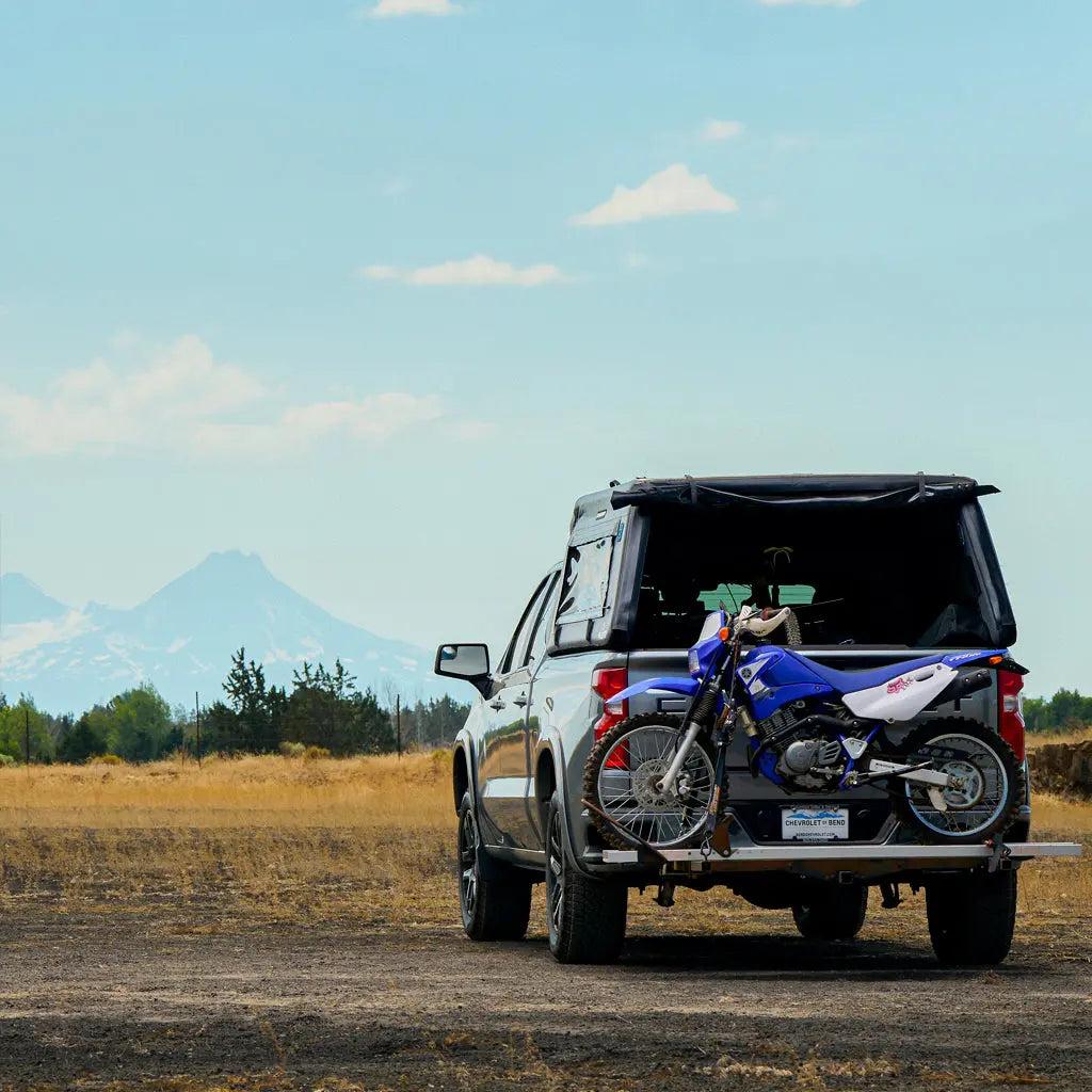 A black pickup truck with a FLATED FULL-STANDARD MR66 topper is parked on a dry field, carrying a blue and white dirt bike on a rear rack. Snow-capped mountains and trees are seen in the background under a blue sky.