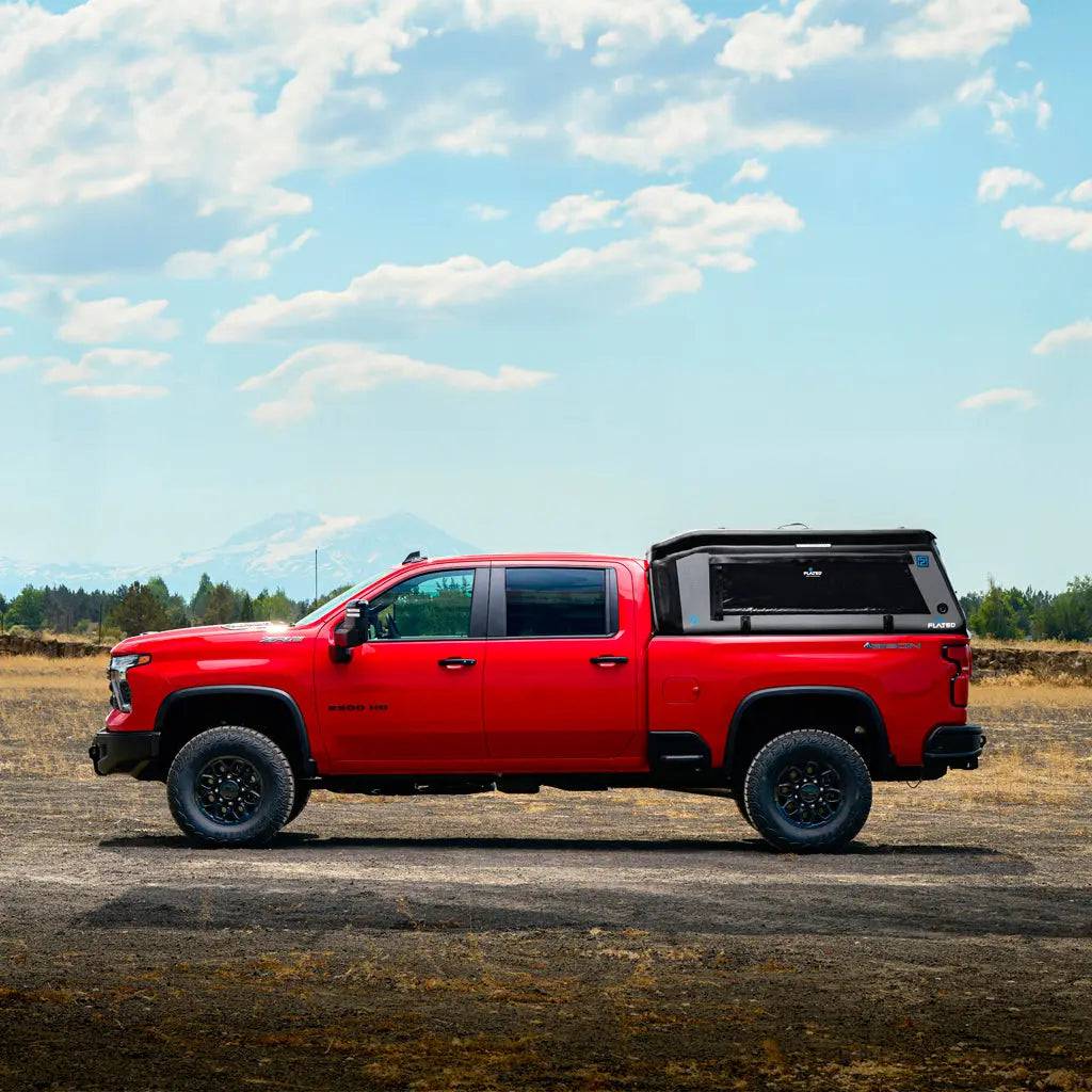A FLATED FULL-STANDARD MR66 bright red pickup with off-road tires and a truck topper is parked on a dirt road in front of fields, trees, and distant mountains under a blue sky with scattered clouds.