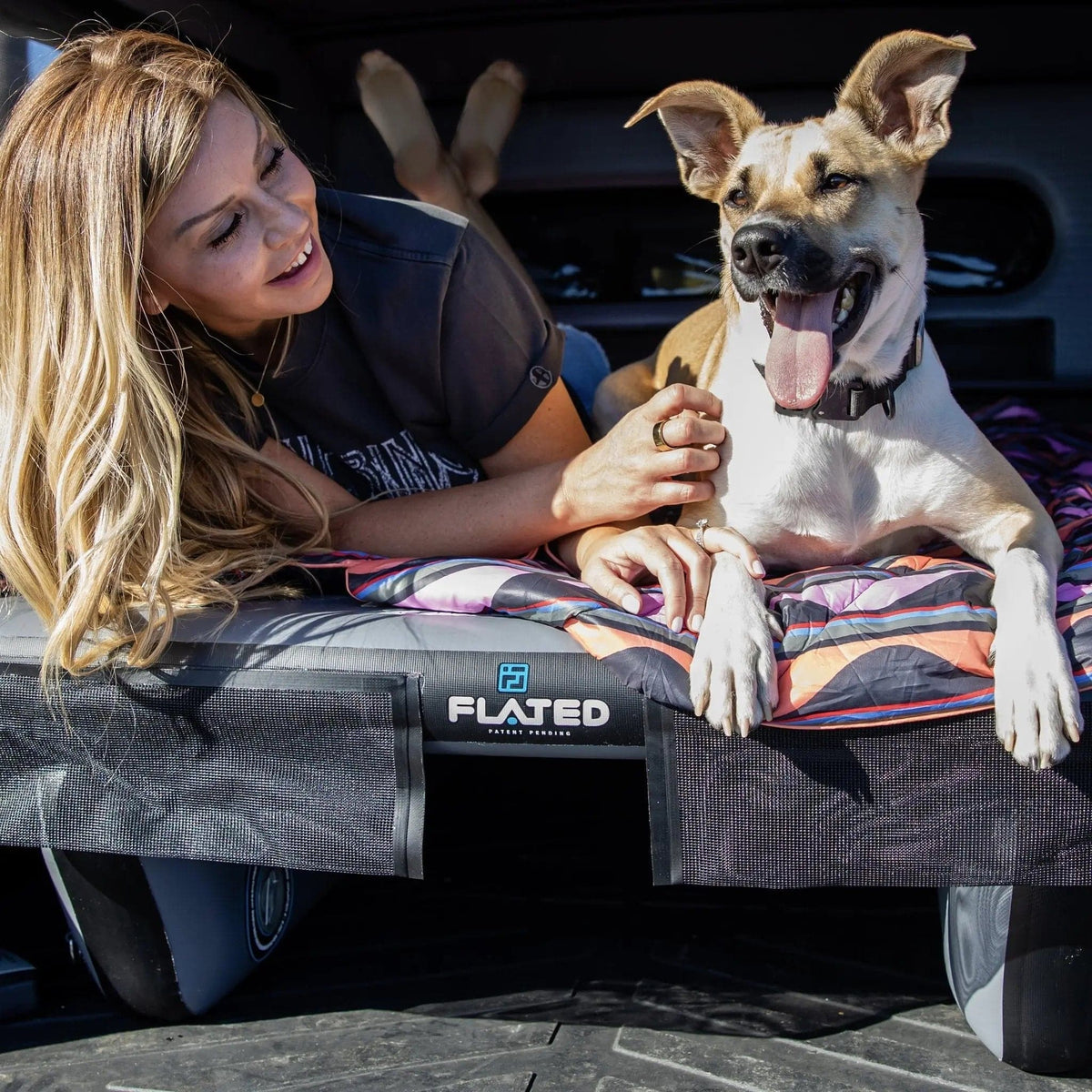 A smiling woman relaxes beside her happy, tan and white dog on a FLATED FULL-SHORT DECK56 inflatable truck bed platform in their adventure rig. Sunlight pours in as her dog, tongue out, enjoys the comfy FULL-SHORT DECK56 with her.