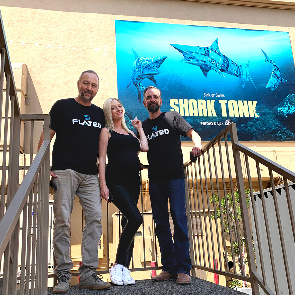 Three people in black FLATED shirts stand on outdoor stairs, smiling, in front of a large Shark Tank TV show billboard featuring mechanical sharks.