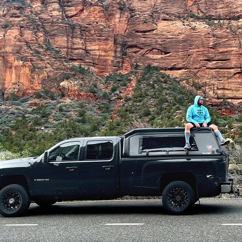 A person in a blue hoodie sits on the roof of a black pickup truck equipped with the FLATED FULL-LONG MR80 topper, parked by a road with red rock cliffs and greenery in the background.