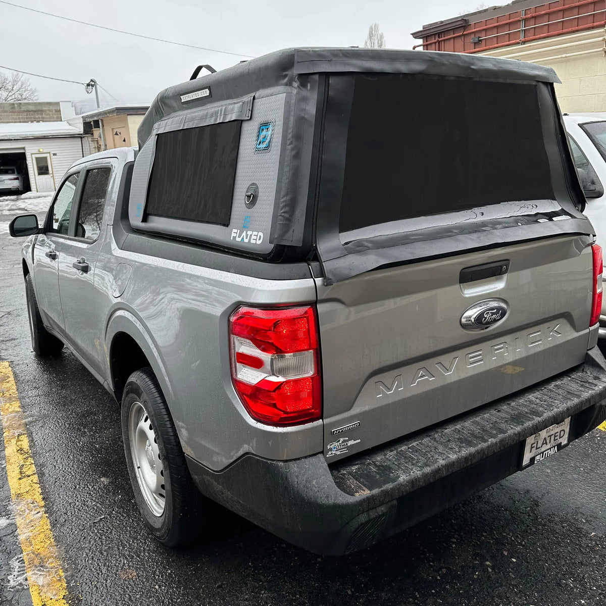 A gray Ford Maverick pickup truck is parked in a lot on a wet, overcast day, featuring the FLATED COMPACT-MAVERICK MR46 inflatable truck shell with a side window covering the bed like a topper.
