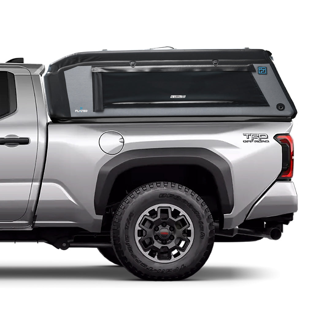 A silver Toyota Tacoma with a 6 ft bed is equipped with FLATED's MID-LONG T24 MR60, a sleek, mid-rise truck topper featuring side windows, off-road tires, and a bold geometric design.