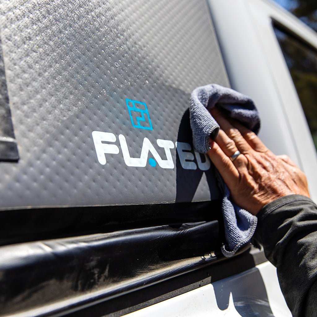 A person uses a gray cloth and FLATED CLEAN & PROTECT to clean a black textured surface with the blue and white FLATED logo on a vehicle, demonstrating proper care for inflatable products.