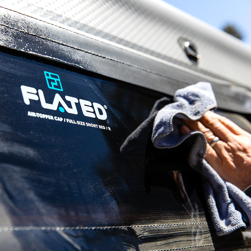 A hand uses a gray cloth and FLATED CLEAN & PROTECT to clean a shiny black surface with the FLATED logo and "AIR-TOPPER CAP / FULL-SIZE SHORT BED | R" text.