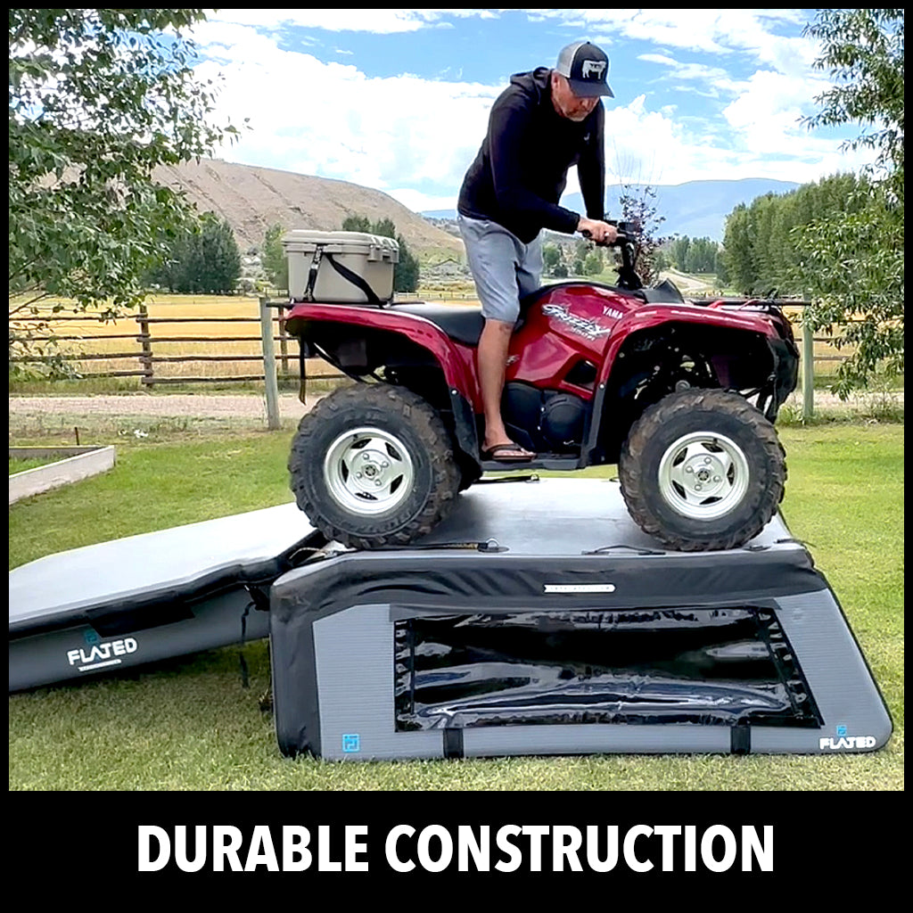 A man on a red ATV drives over a black inflatable platform outdoors, demonstrating its strength. The background shows trees, grass, and distant hills under a blue sky. Text below reads: DURABLE CONSTRUCTION.