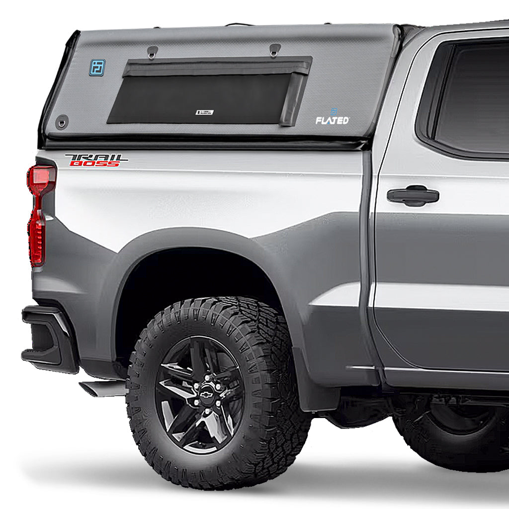 A silver pickup truck with large off-road tires has a black, boxy FLATED FULL-SHORT CAP56 Air-Topper truck bed camper, featuring a window and the FLATED brand name on the side.