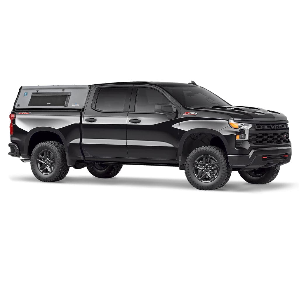 A silver pickup truck with large off-road tires has a black, boxy FLATED FULL-SHORT CAP56 Air-Topper truck bed camper, featuring a window and the FLATED brand name on the side.